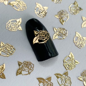 May include: Gold metal rose nail art embellishments. The rose shapes are delicate and intricate. The nail art is on a black nail polish background.