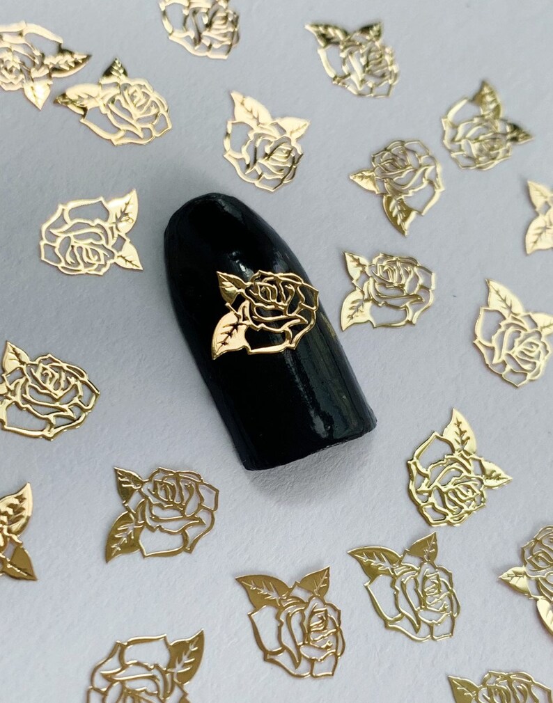 May include: Gold metal rose nail art embellishments. The rose shapes are delicate and intricate. The nail art is on a black nail polish background.