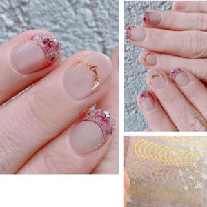 May include: Close-up of a hand with pink and white nail polish and gold glitter accents. The nails have a half-moon design with gold glitter on the bottom half of each nail. The image also shows a sheet of gold crescent moon nail stickers.