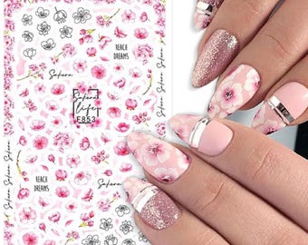 May include: Nail art stickers featuring pink floral designs and the text "Reach Dreams" and "Safara". The stickers are on a white background. Some nails are painted with pink and glitter polish, and floral designs.