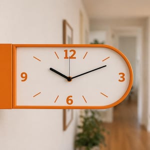 May include: An orange and white wall clock with a rectangular base and an oval clock face. The clock has black hands and orange numerals at the 12, 3, 6, and 9 positions. The clock is mounted on a white wall.