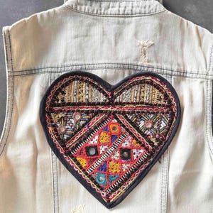 Könnte beinhalten: Eine ärmellose Jeansweste in hellem Waschton, mit einem großen, gestickten Herzaufnäher auf dem Rücken. Das Herzdesign enthält verschiedene Farben und Muster, darunter Rot, Gelb und Blau, mit Spiegelakzenten.