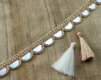 May include: Gold trim with mirrored scallops and two tassels, one white and one beige, on a brown fabric background.