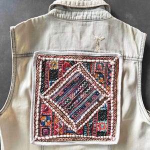 May include: A light-colored denim vest with a colorful, embroidered patch on the back. The patch features a geometric design with red, blue, and white accents, and is framed by a white border.