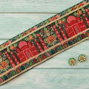 以下が含まれることがあります： 緑色の生地のトリムに、金糸の刺繍が施され、ピンクと赤のタージ・マハルがデザインされています。トリムは薄い青色の木製の表面に置かれています。