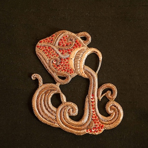 May include: A gold-coloured metal embroidery of the astrological sign Aquarius. The design features a stylised water pitcher pouring water, with red beads representing the water.