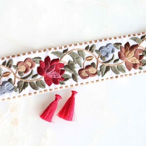 Pode incluir: Fita bordada branca com um design floral em tons de vermelho, castanho, azul e verde. Duas pequenas borlas vermelhas estão colocadas por baixo. Ideal para costura ou artesanato.