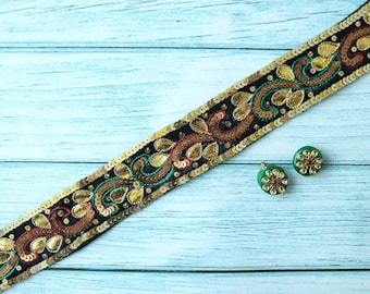 May include: Black sequined trim with gold and copper floral designs and teal accents. Two green earrings with gold and red accents are also visible. The trim and earrings are on a light blue wooden surface.