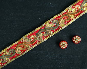 Puede incluir: Una tira de tela roja con bordes de lentejuelas doradas y bordados dorados intrincados con detalles en hilo verde azulado, acompañada de dos pendientes redondos rojos a juego con un diseño floral central y pequeñas piedras transparentes.
