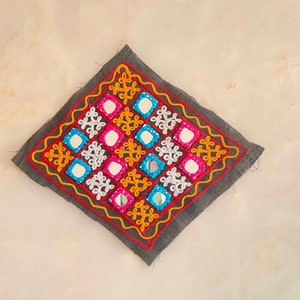 Puede incluir: Un cuadrado de tela gris con un diseño bordado de colores que incluye pequeños espejos. El diseño incluye hilo amarillo, naranja, rosa, azul y blanco.