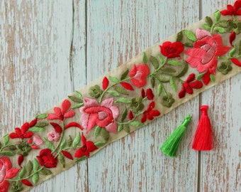 May include: A beige fabric trim with embroidered pink and red flowers and green leaves. The trim is on a light blue wooden surface.