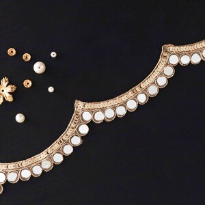 May include: Gold trim with round mirrored embellishments on a black fabric background. There are also small gold flower-shaped embellishments and white beads scattered on the fabric.