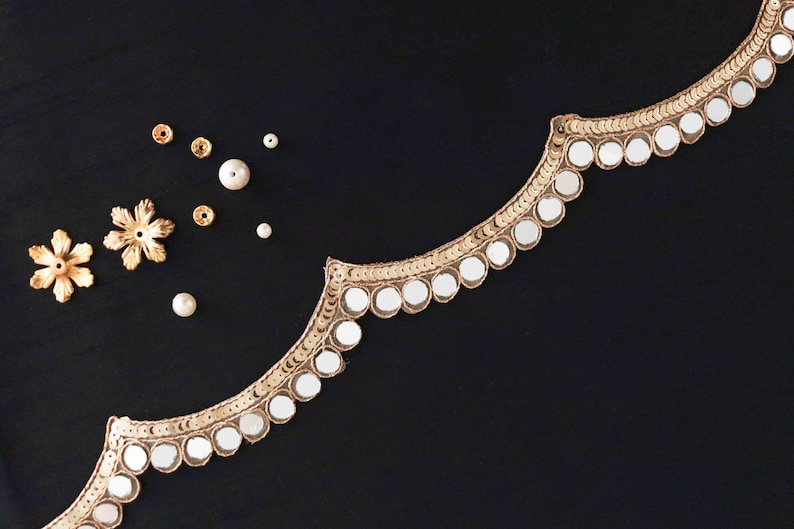 May include: Gold trim with round mirrored embellishments on a black fabric background. There are also small gold flower-shaped embellishments and white beads scattered on the fabric.