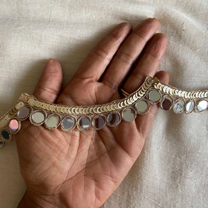 May include: Gold trim with round, mirrored sequins. The trim is shown on a hand.