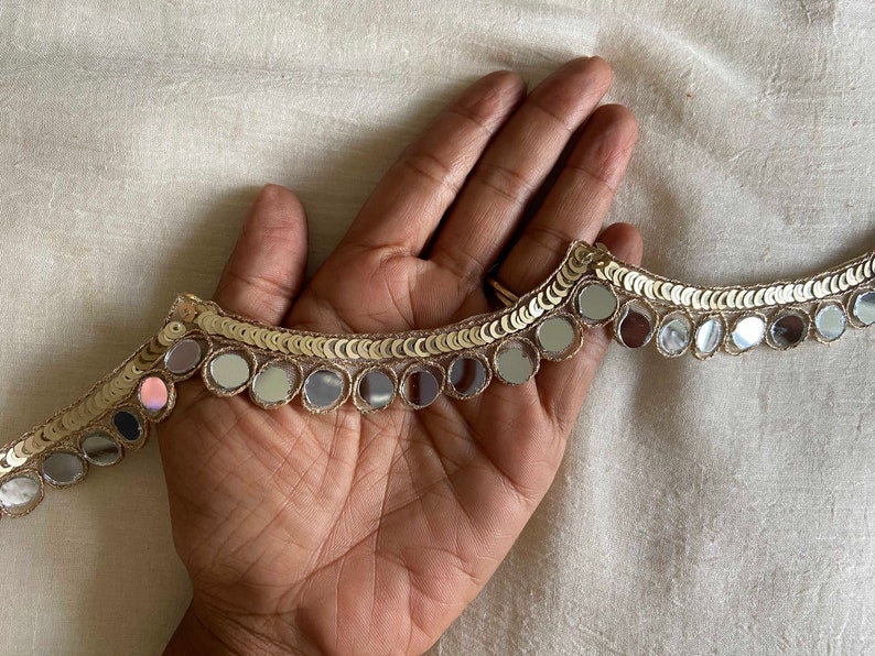 May include: Gold trim with round, mirrored sequins. The trim is shown on a hand.