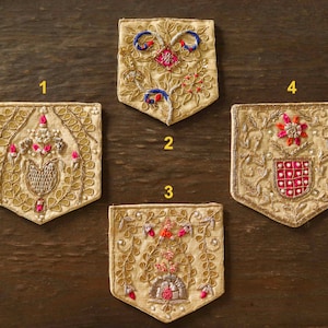 Puede incluir: Cuatro parches de tela bordados con diseños florales en tonos rosa, rojo, azul y dorado. Los parches son beige y tienen un borde festoneado.
