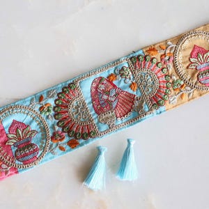 May include: A decorative fabric trim featuring a repeating pattern of embroidered circular motifs, stylized figures, and floral elements in shades of pink, light blue, and gold, accompanied by two small, light blue thread tassels.