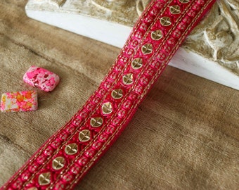 Pode incluir: Bordado rosa e dourado com um padrão repetido de pequenos círculos dourados e um círculo dourado maior no centro de cada padrão. O bordado está sobre uma superfície marrom.