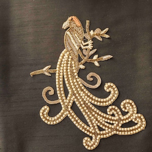 May include: A gold and white embroidered peacock design with a long tail of pearls. The peacock is perched on a branch with leaves.