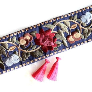 Peut inclure: Garniture florale brodée bleu marine avec des fleurs rouges, marron et bleues, des feuilles vertes et des détails dorés. Deux pompons roses vifs sont présents. Idéal pour divers projets créatifs.