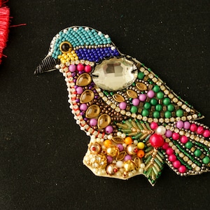 May include: A colourful beaded bird applique with a large clear rhinestone in the centre. The bird is made with various colours of beads, including blue, green, pink, gold, and white.
