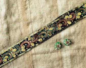 May include: A black decorative ribbon with gold, teal, and maroon sequins and floral designs. Two green floral earrings with red and white accents are also visible on a beige fabric background.