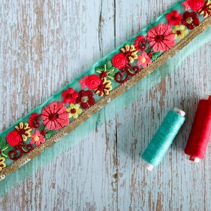 May include: A turquoise blue fabric trim with gold sequins and red, pink, and yellow embroidered flowers. Two spools of thread, one turquoise blue and one red, are on a whitewashed wooden surface.