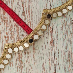 May include: Gold trim with round mirror embellishments. The trim is on a light wood background with a red measuring tape.