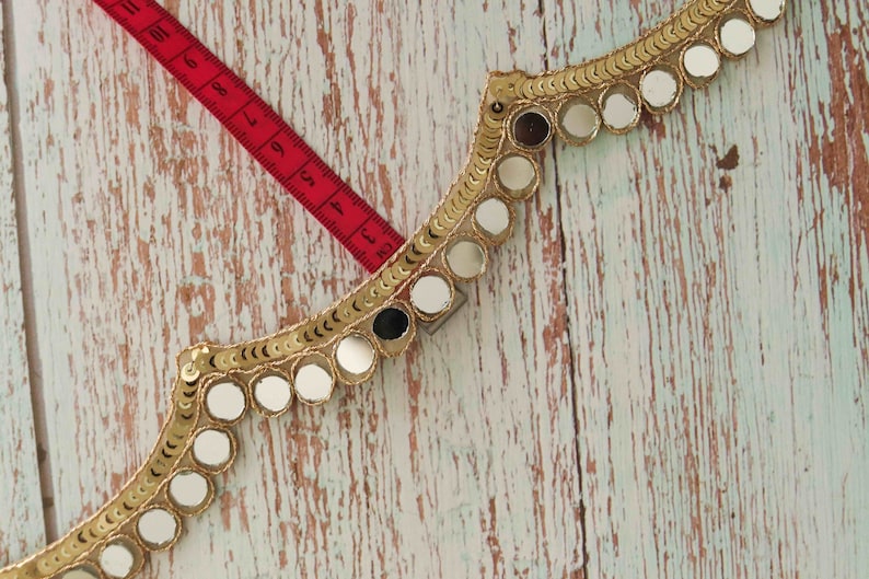 May include: Gold trim with round mirror embellishments. The trim is on a light wood background with a red measuring tape.