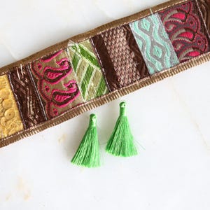 May include: A decorative textile strip with a variety of patterned fabric sections in orange, yellow, brown, green, and teal. Two bright green tassels are below the strip. The strip has a gold border and is on a white surface.