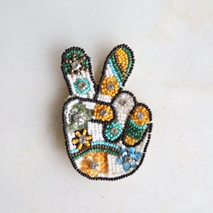 May include: A beaded peace sign pin with a white base, outlined in black beads. The pin features colorful floral and geometric patterns in yellow, turquoise, and green, with silver accents. The pin is approximately 3 inches tall.