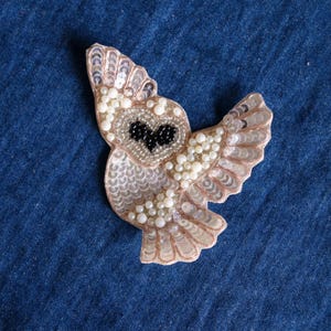May include: An owl-shaped patch featuring intricate beadwork and sequins. The owl has a heart-shaped face outlined in silver beads with black bead eyes, and wings covered in shimmering pale pink sequins and small white pearl-like beads.
