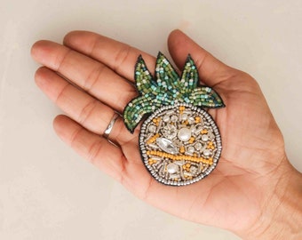 Puede incluir: Un broche de piña con cuentas, con hojas verdes y amarillas, cuentas blancas y amarillas y un centro plateado.
