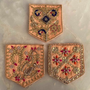 Puede incluir: Tres parches de tela bordados con intrincados diseños florales y geométricos. Los parches son beige con bordados dorados, rosados y azules.