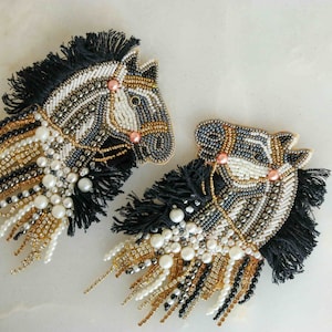 Puede incluir: Dos broches de cabeza de caballo con cuentas de cebra en blanco y negro con flecos y detalles de perlas.