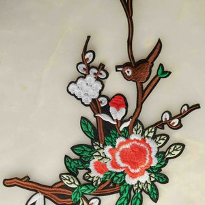 May include: Embroidered iron-on patch featuring a brown bird perched on a branch with green leaves, white blossoms, and a red flower.