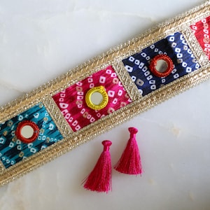 May include: A decorative fabric trim featuring a series of square panels with tie-dye patterns in teal, magenta, navy, and red, each centred with a small circular mirror. The trim is bordered by gold metallic embroidery and includes two pink tassels.