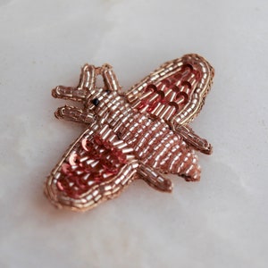 May include: A rose gold beaded and sequined bee applique. The bee is made of small, round beads and sequins in a rose gold colour. The bee has a detailed body and wings.