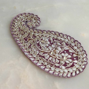 May include: A decorative paisley-shaped embellishment with a deep purple base. The edges are outlined with silver-coloured beads, and the interior features intricate floral patterns. The piece is likely used for embellishing clothing or accessories.