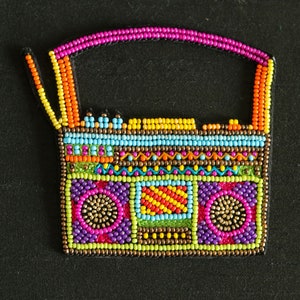 以下が含まれることがあります： 紫、緑、黄色のデザインのカラフルなビーズ製のブームボックス。ブームボックスは、小さなカラフルなビーズを縫い合わせて、詳細な画像を作成しています。