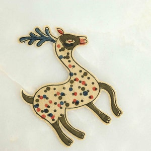 Puede incluir: Una ilustración caprichosa de un ciervo manchado con una cornamenta azul y verde, pintada en tonos de marrón, rojo y azul.