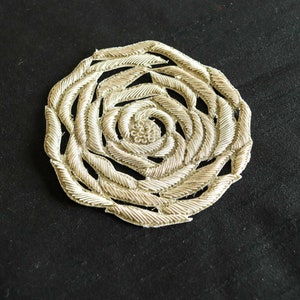 Könnte beinhalten: Eine silberne gestickte Rosenapplikation in runder Form. Die Rose besteht aus einem zarten, fadenartigen Material und hat ein erhabenes, strukturiertes Design.