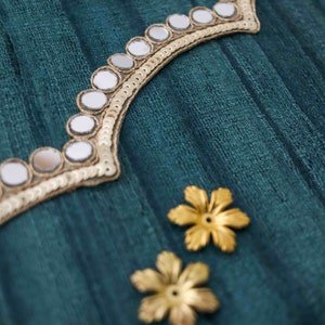 May include: Teal fabric with gold trim and mirrored embellishments. Two gold flower-shaped embellishments are also visible.