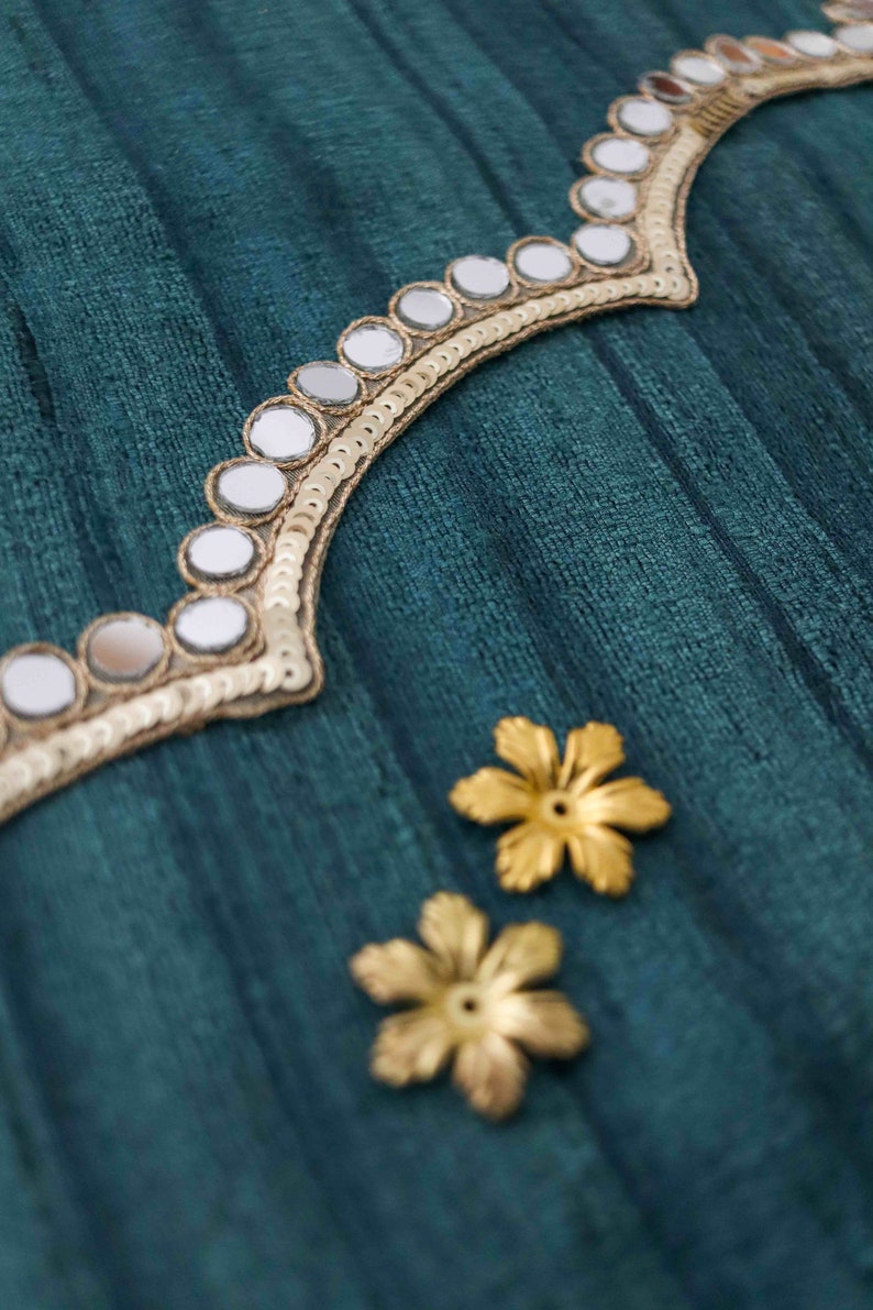 May include: Teal fabric with gold trim and mirrored embellishments. Two gold flower-shaped embellishments are also visible.