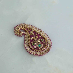 Puede incluir: Un parche de tela de color rosa en forma de paisley con adornos dorados y verdes. El parche está decorado con un diseño floral y pequeñas piedras brillantes.