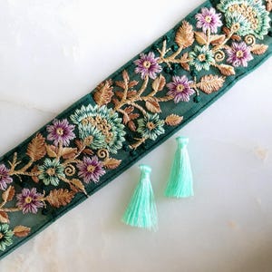 May include: A strip of dark green mesh fabric featuring intricate floral embroidery in shades of purple, light green, and tan, accompanied by two small, light mint green tassels positioned separately from the embroidered fabric trim.