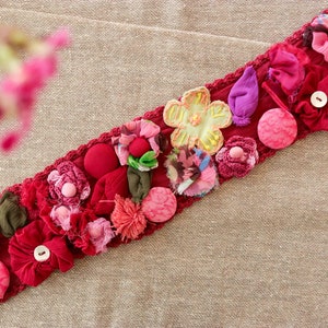 Könnte beinhalten: Ein roter Stoffbesatz mit verschiedenen Stoffblumen und Knöpfen. Der Besatz zeigt rosa, gelbe, grüne und violette Stoffblumen sowie rote und rosa Knöpfe.
