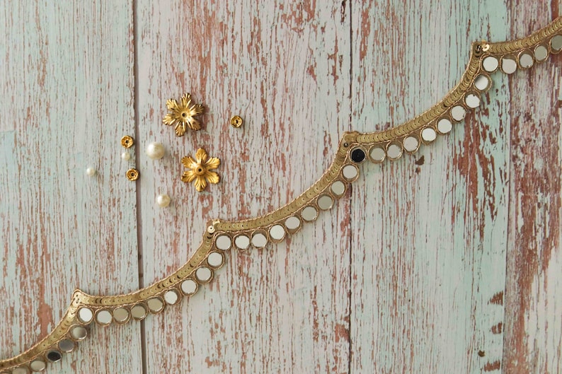 May include: Gold trim with round mirrored accents on a light blue wood background. There are also small gold and white embellishments scattered around the trim.