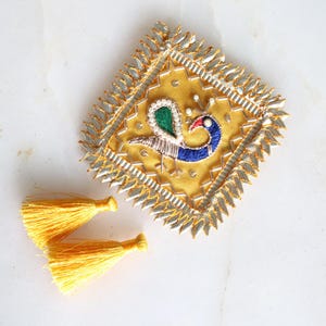 May include: A square, yellow fabric embellishment with a peacock design, featuring blue, green, and red embroidery. The edges are trimmed with silver and gold detailing. Two yellow tassels are placed below the embellishment.
