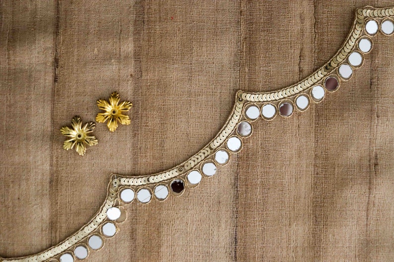 May include: A gold trim with round mirror embellishments on a brown fabric background. Two gold star-shaped embellishments are also visible.
